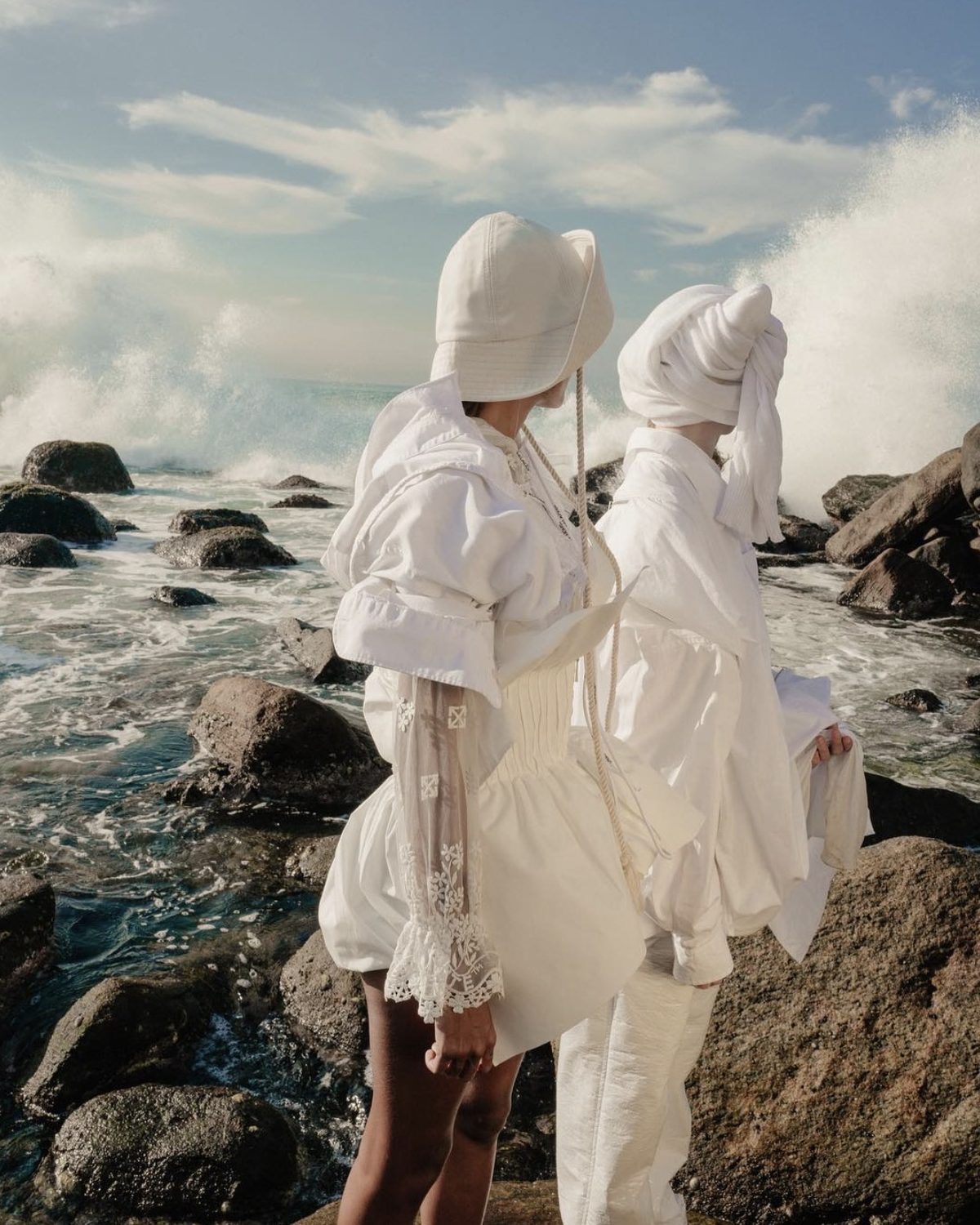 Two figures dressed in white on the rocks as waves break behind them